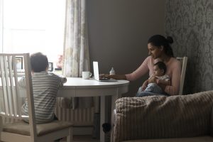 Young mother sitting at the table at home. She is holding her baby son whilst using a laptop. Her other son is sitting with his back to the camera, colouring in at the table.