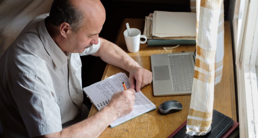 Older man sitting at desk and writing in notebook.