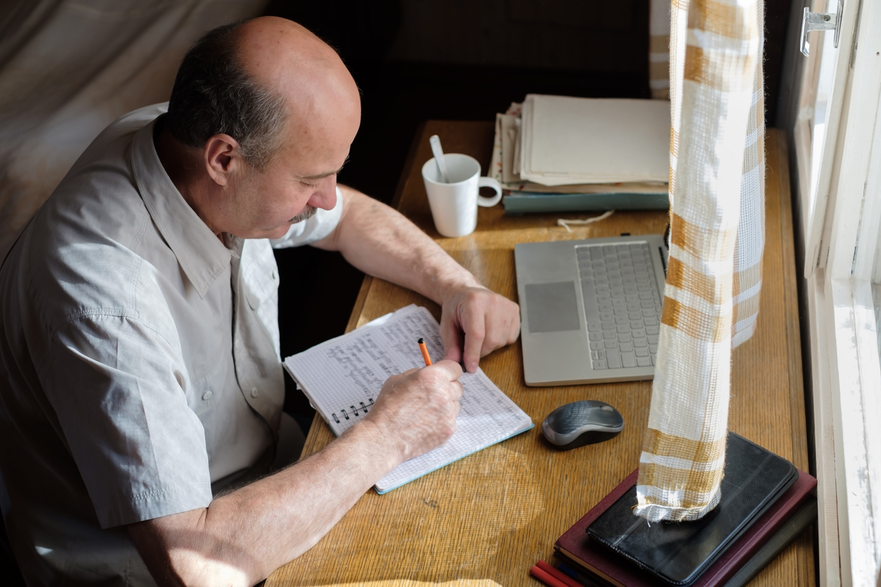 Older man sitting at desk and writing in notebook.