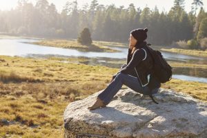 Young woman wearing backpack sitting on rock in forested area overlooking water.