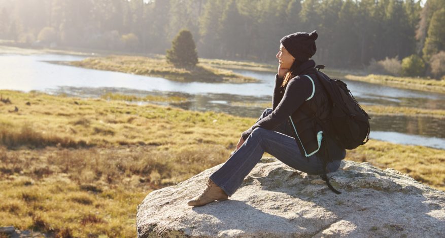 Young woman wearing backpack sitting on rock in forested area overlooking water.