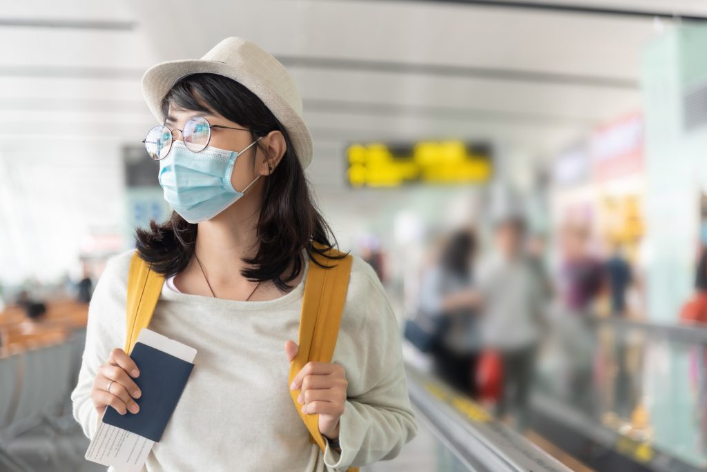 Happy Asian woman wear protective face mask and eyeglasses walking in international airport terminal during virus pandemic. Smiling Young Female traveler with yellow backpack at the departure hall