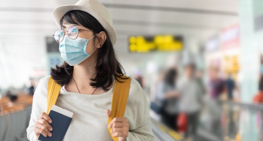 Happy Asian woman wear protective face mask and eyeglasses walking in international airport terminal during virus pandemic. Smiling Young Female traveler with yellow backpack at the departure hall