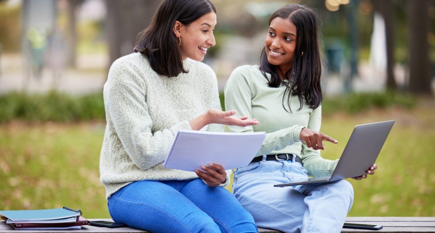 Two women sitting on bench talking and looking at laptop