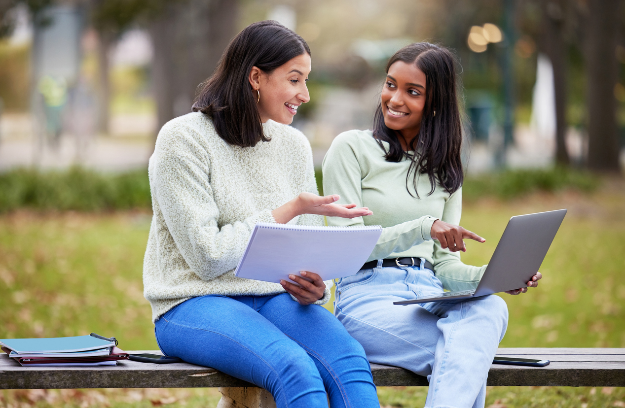 Two women sitting on bench talking and looking at laptop