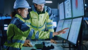 Two engineers wearing white safety hats and yellow jackets pressing on touch-screen computer