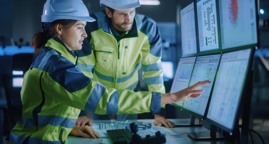 Two engineers wearing white safety hats and yellow jackets pressing on touch-screen computer