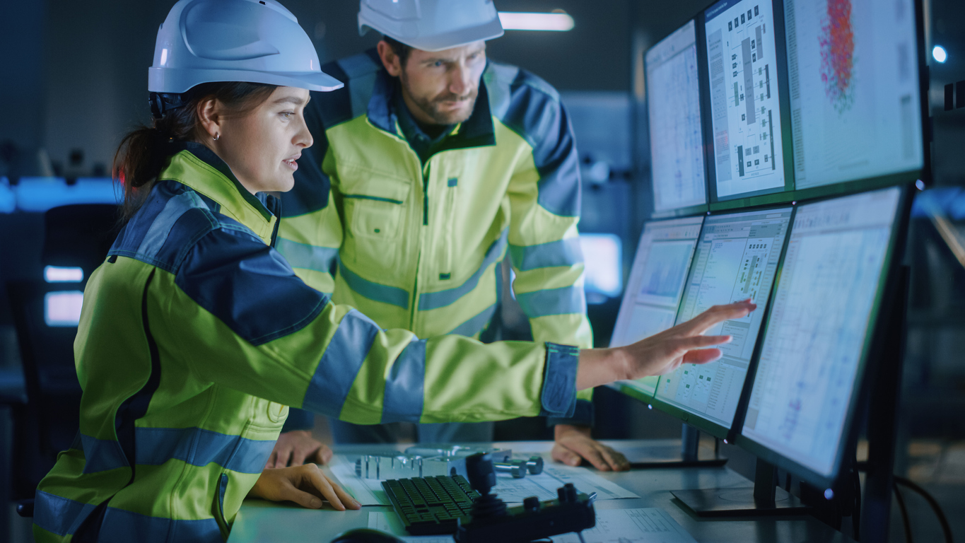 Two engineers wearing white safety hats and yellow jackets pressing on touch-screen computer