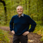 Adriano along a hiking path surrounded by greenery, wearing a navy blue sweater and a blue shirt collar. He has his hands on his hips and he smiles with his teeth, facing the camera.