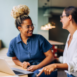 Two young women smiling and talking while working together at a laptop