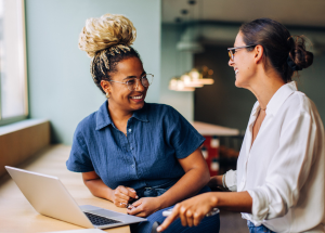 Two young women smiling and talking while working together at a laptop